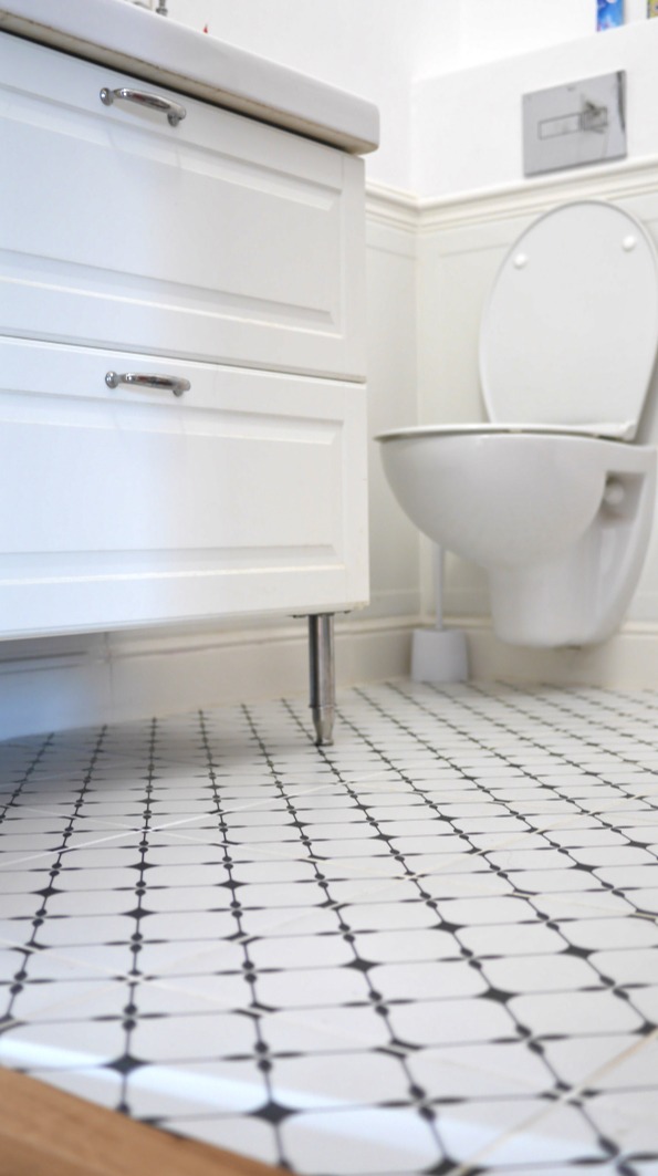 Biała szafka pod umywalkę z chromowanymi uchwytami i nóżkami na tle toalety i białych paneli ściennych, wzorzysta biało-czarna podłoga z płytek.