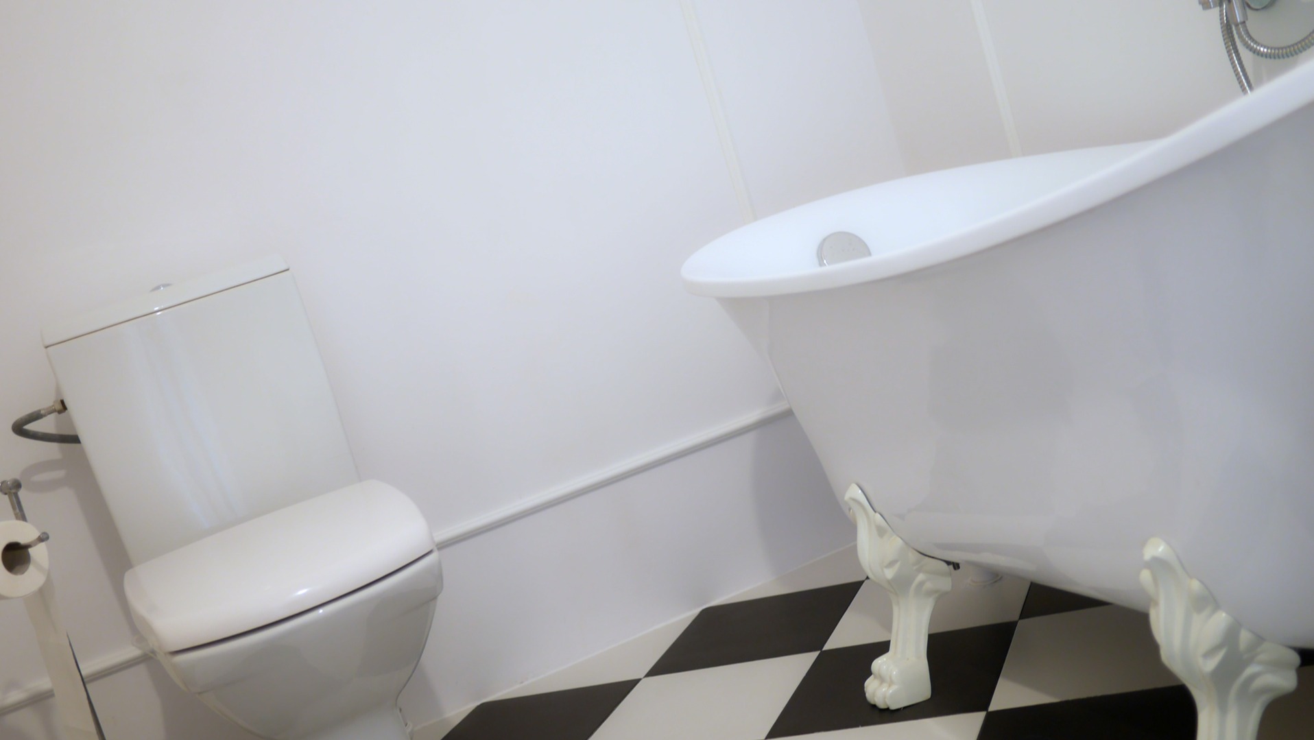 Biała wolnostojąca wanna na ozdobnych nóżkach oraz toaleta w minimalistycznej łazience z czarno-białą podłogą w szachownicę.