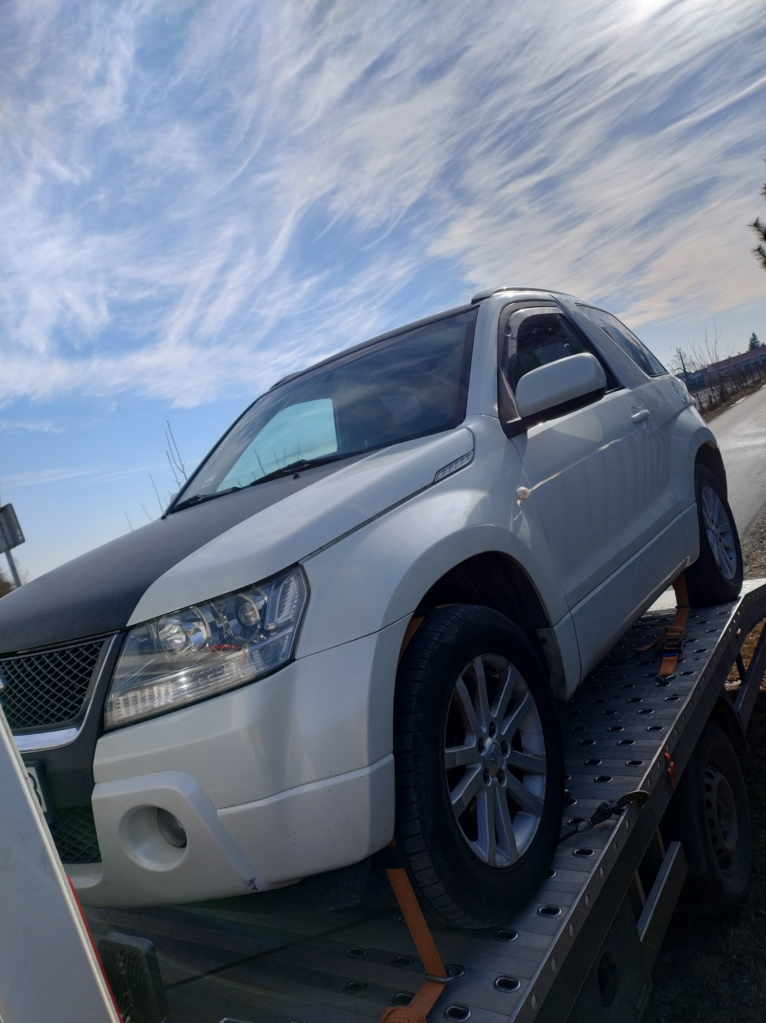 Biały samochód Suzuki Grand Vitara z czarną maską, zabezpieczony pasami, transportowany na platformie pomocy drogowej w słoneczny dzień.