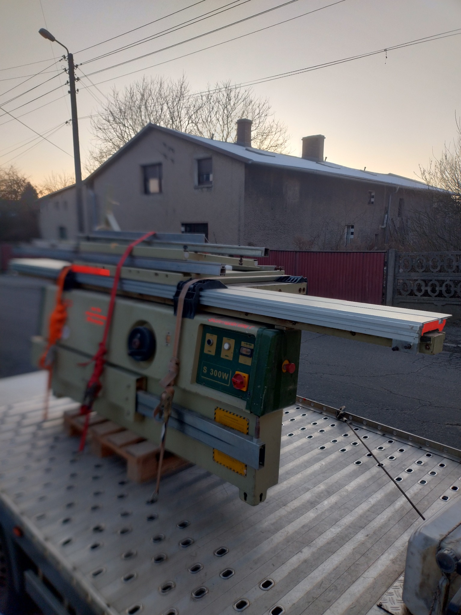 Maszyna stolarska S 300W na palecie, zabezpieczona pasami, transportowana na platformie lawety na tle budynku wczesnym rankiem.