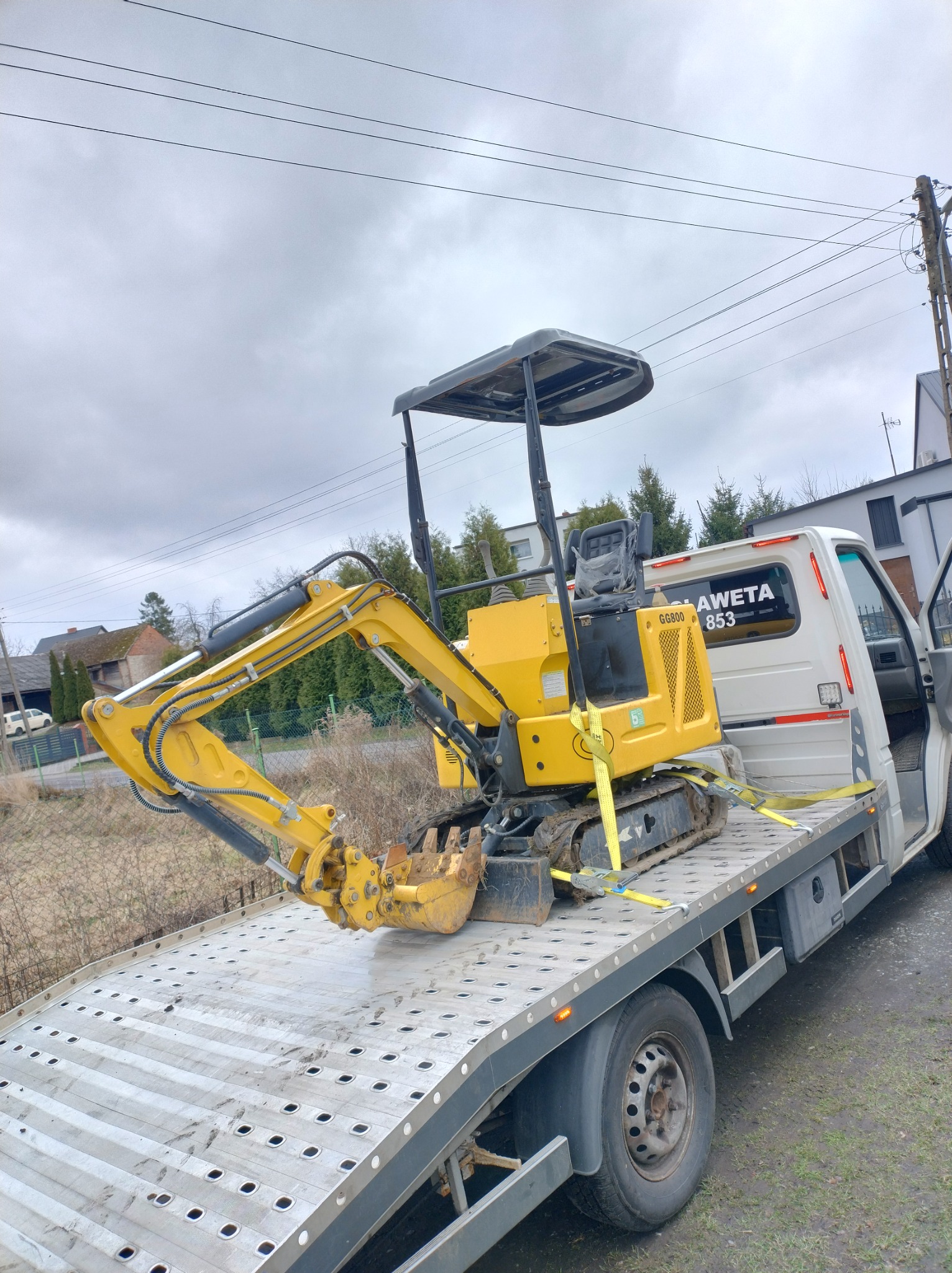 Żółta mini koparka GG800 zabezpieczona pasami na platformie pojazdu z napisem na szybie. Transport maszyny budowlanej w pochmurny dzień.
