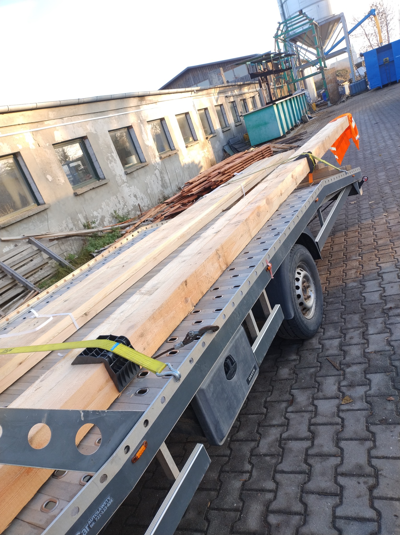 Drewno zabezpieczone pasami na przyczepie transportowej z pomarańczową flagą ostrzegawczą, w tle budynek i silos.