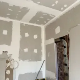 Wnętrze w trakcie prac wykończeniowych: ściany z płyt gipsowo-kartonowych, częściowo pokryte szpachlą, widoczne instalacje elektryczne, żółta skrzynka z narzędziami w rogu pomieszczenia.