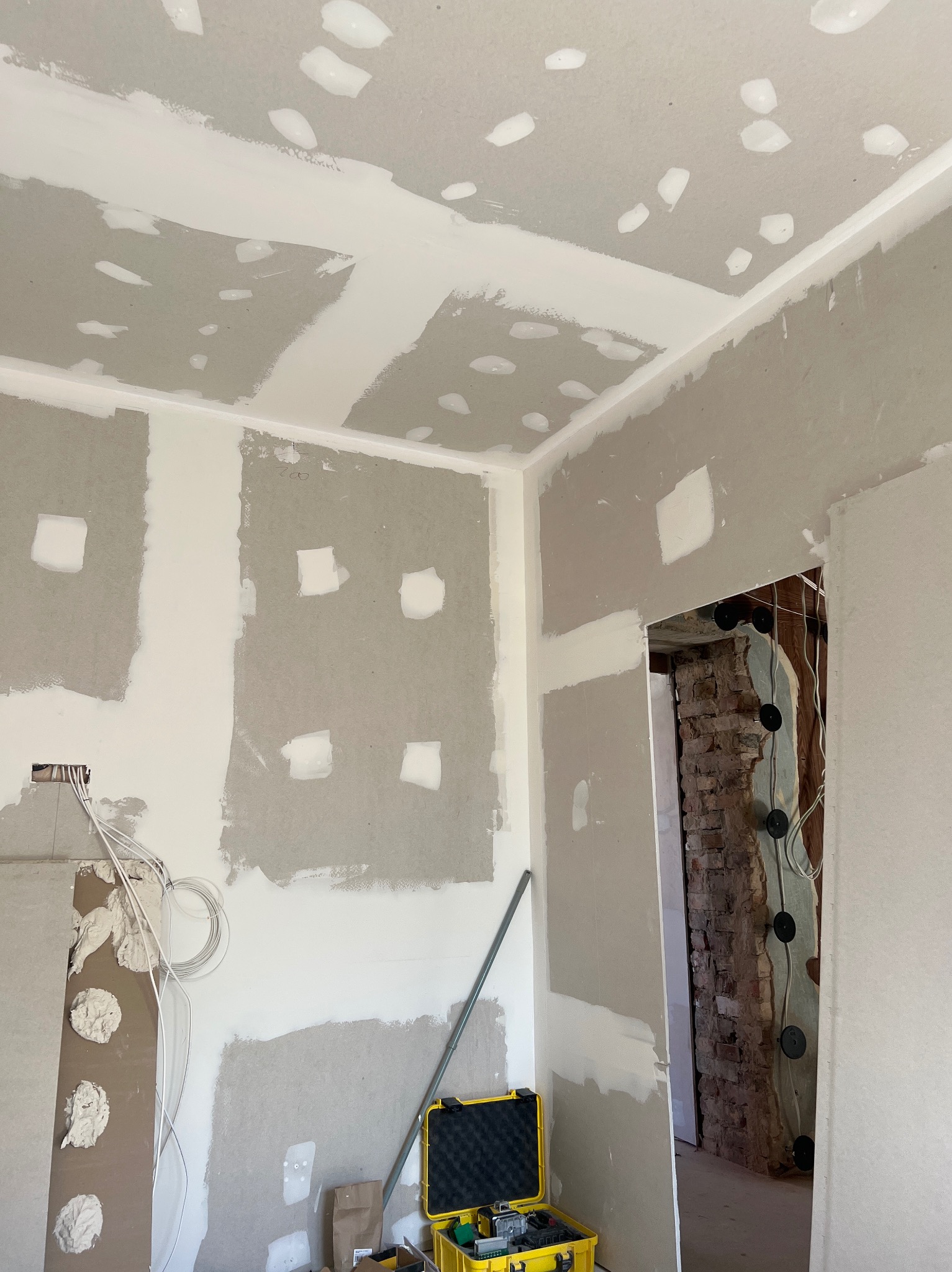 Wnętrze w trakcie prac wykończeniowych: ściany z płyt gipsowo-kartonowych, częściowo pokryte szpachlą, widoczne instalacje elektryczne, żółta skrzynka z narzędziami w rogu pomieszczenia.