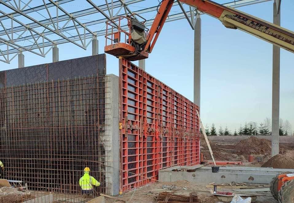 Montaż szalunku na fundamencie budynku przemysłowego, widoczna siatka zbrojeniowa i pracownik w żółtym kasku, w tle stalowa konstrukcja dachu i podnośnik koszowy.