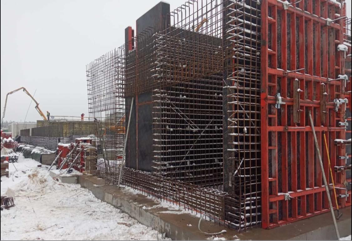 Zimowy widok na plac budowy z szalunkami i zbrojeniami fundamentów, z pompą do betonu w tle i śniegiem na ziemi.