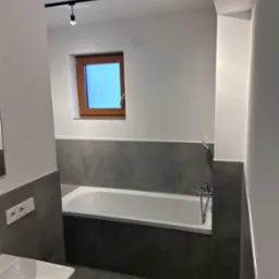 Wnętrze łazienki po remoncie: biała wanna obudowana szarymi płytkami, szara podłoga, okno z drewnianą ramą, biała umywalka i nowoczesne oświetlenie punktowe na suficie.