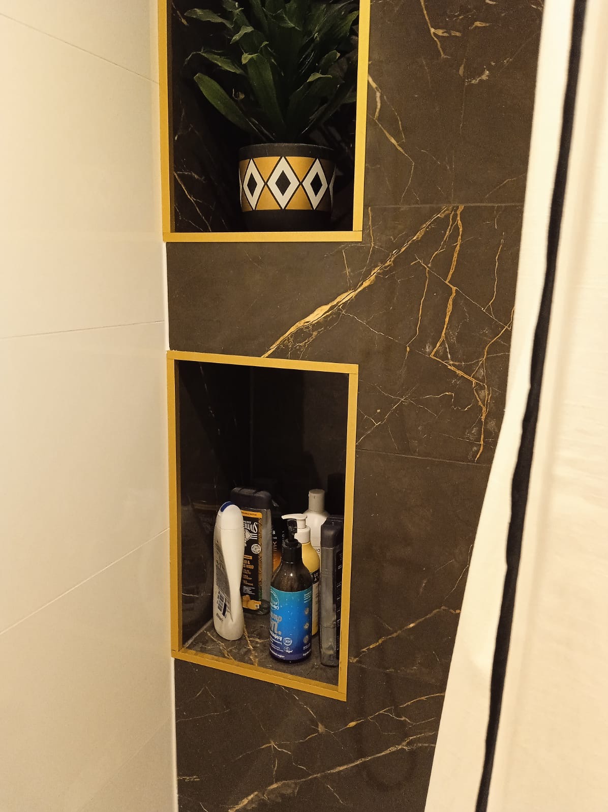 Wnęki w łazience wykończone czarnym marmurem z złotymi żyłami, jedna z rośliną w ozdobnej doniczce, druga z kosmetykami, obramowane złotymi listwami.