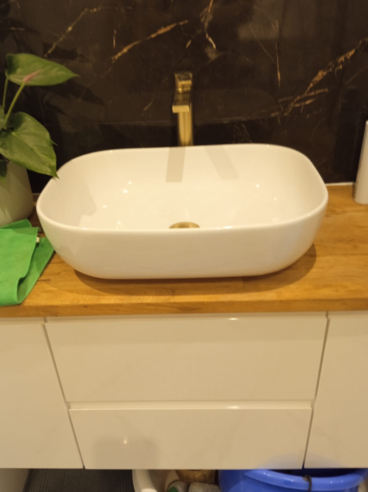 Nowoczesna, prostokątna umywalka nablatowa z złotą armaturą na drewnianym blacie i białych szafkach, na tle ściany z ciemnego marmuru z jasnymi żyłami.