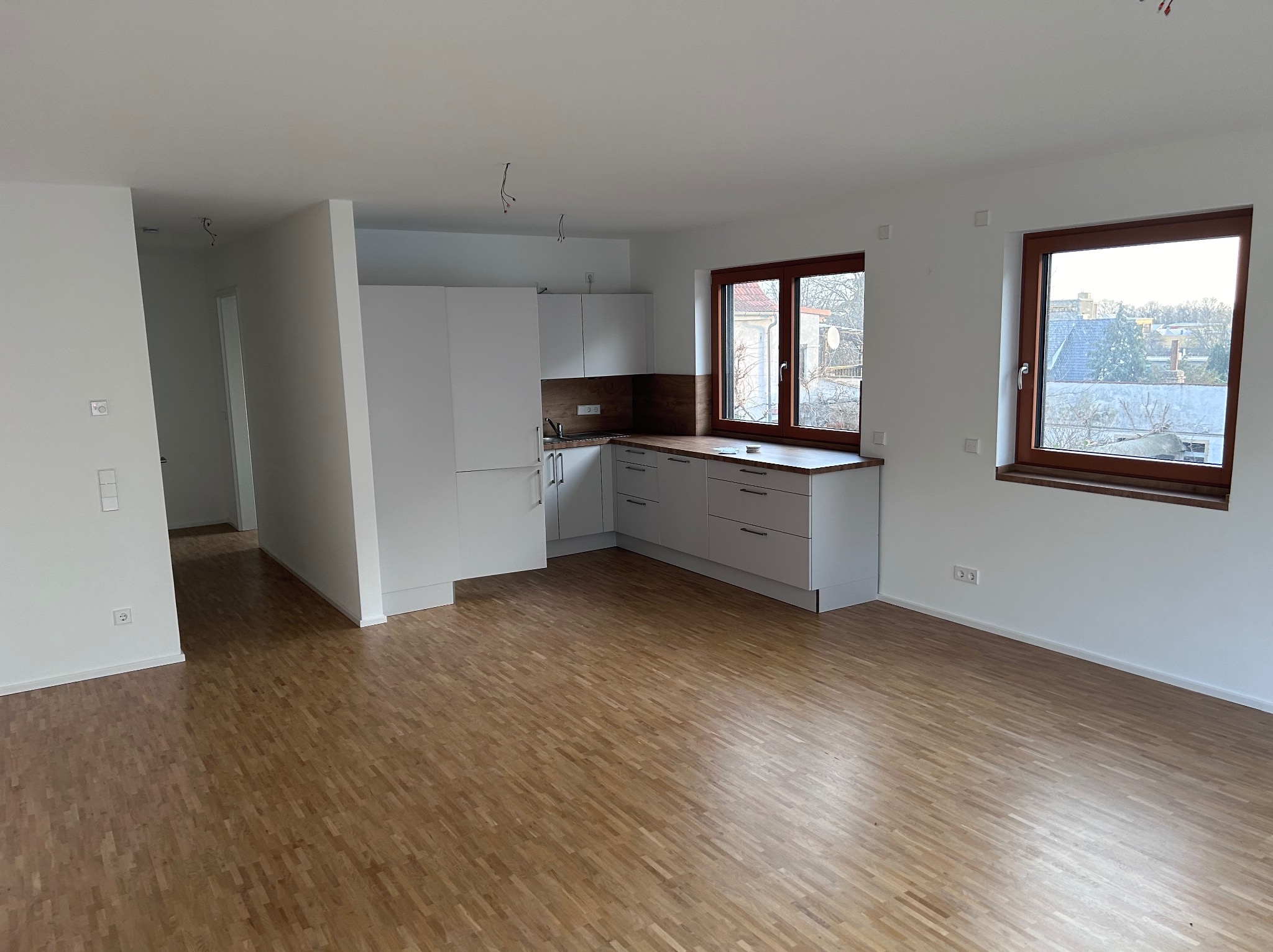 Nowoczesne, jasne wnętrze z drewnianą podłogą, białą kuchnią w zabudowie i dwoma oknami z brązowymi ramami. Widoczne wystające kable z sufitu sugerują niedokończone prace elektryczne.