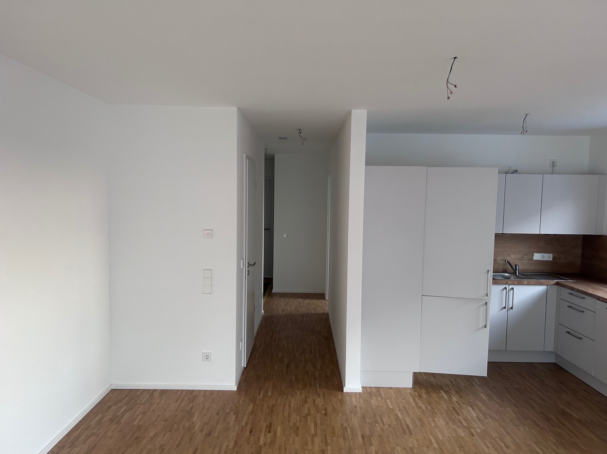 Wnętrze pustego mieszkania po remoncie: białe ściany, drewnopodobne panele podłogowe, widoczna część kuchni z białymi szafkami i drewnianym blatem, oraz fragment korytarza z drzwiami.