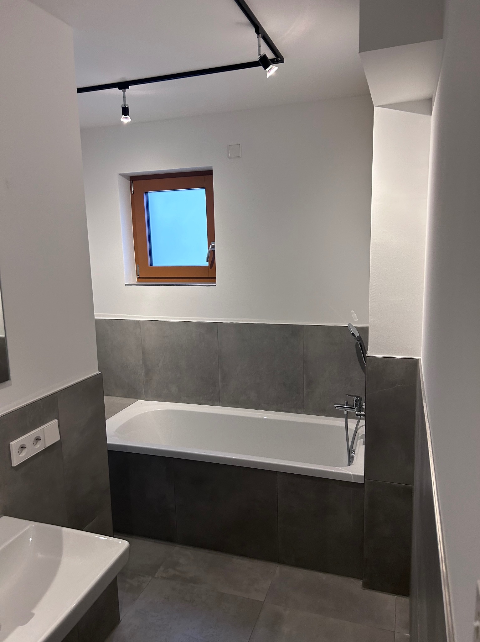 Wnętrze łazienki po remoncie: biała wanna obudowana szarymi płytkami, szara podłoga, okno z drewnianą ramą, biała umywalka i nowoczesne oświetlenie punktowe na suficie.