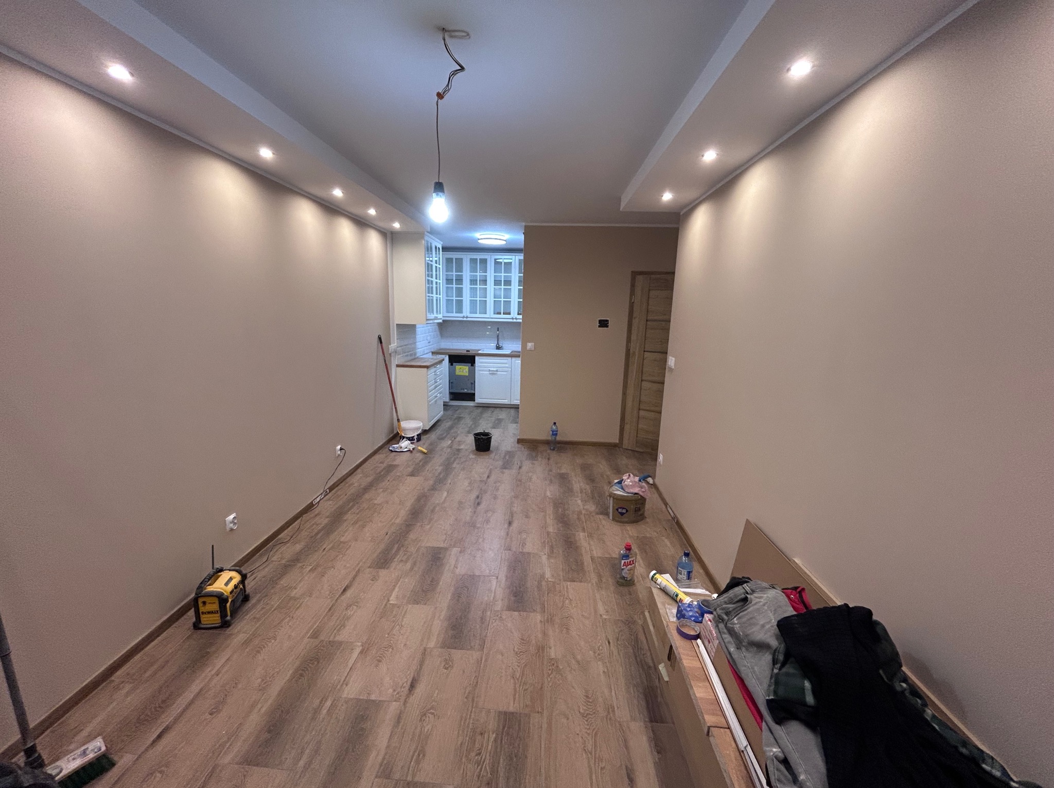 Jasne wnętrze po remoncie: świeżo pomalowane ściany w odcieniach beżu, panele podłogowe imitujące drewno, sufit z oświetleniem punktowym, widok na kuchnię.