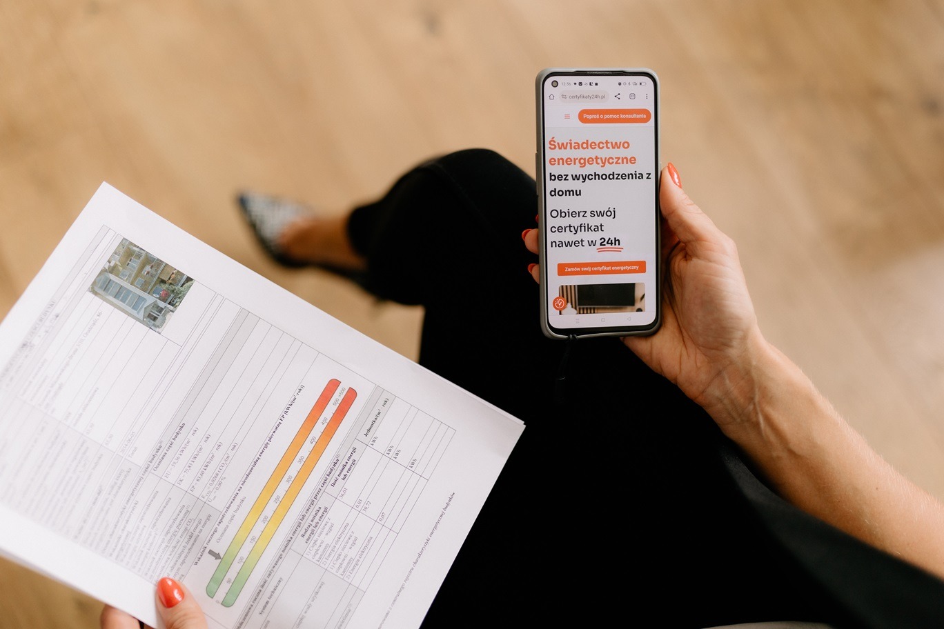 Osoba trzymająca telefon z wyświetloną stroną internetową oferującą certyfikaty energetyczne w 24h, obok leży dokument z danymi charakterystyki energetycznej budynku oraz zdjęciem budynku w rogu.