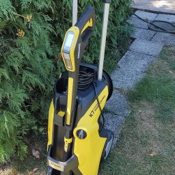 CleanViolFresh - Żółto-czarna myjka ciśnieniowa Karcher K7 Smart Control, stojąca na trawie obok zielonego żywopłotu i kamiennej ścieżki.