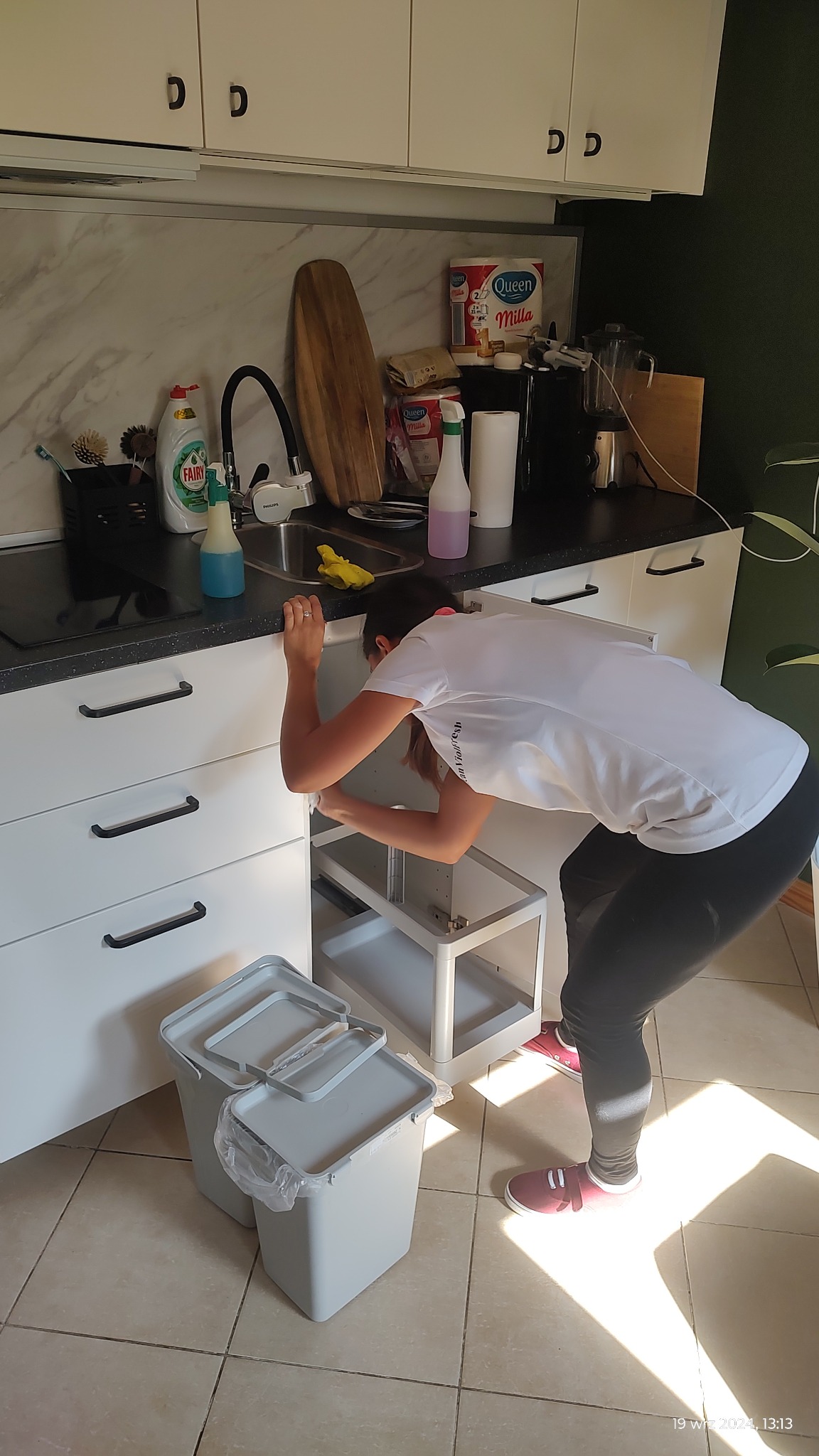 Kobieta w białej koszulce czyści wnętrze szafki pod zlewem w kuchni, obok stoją dwa pojemniki na śmieci z szarymi pokrywkami, na blacie widoczne środki czystości i akcesoria kuchenne.