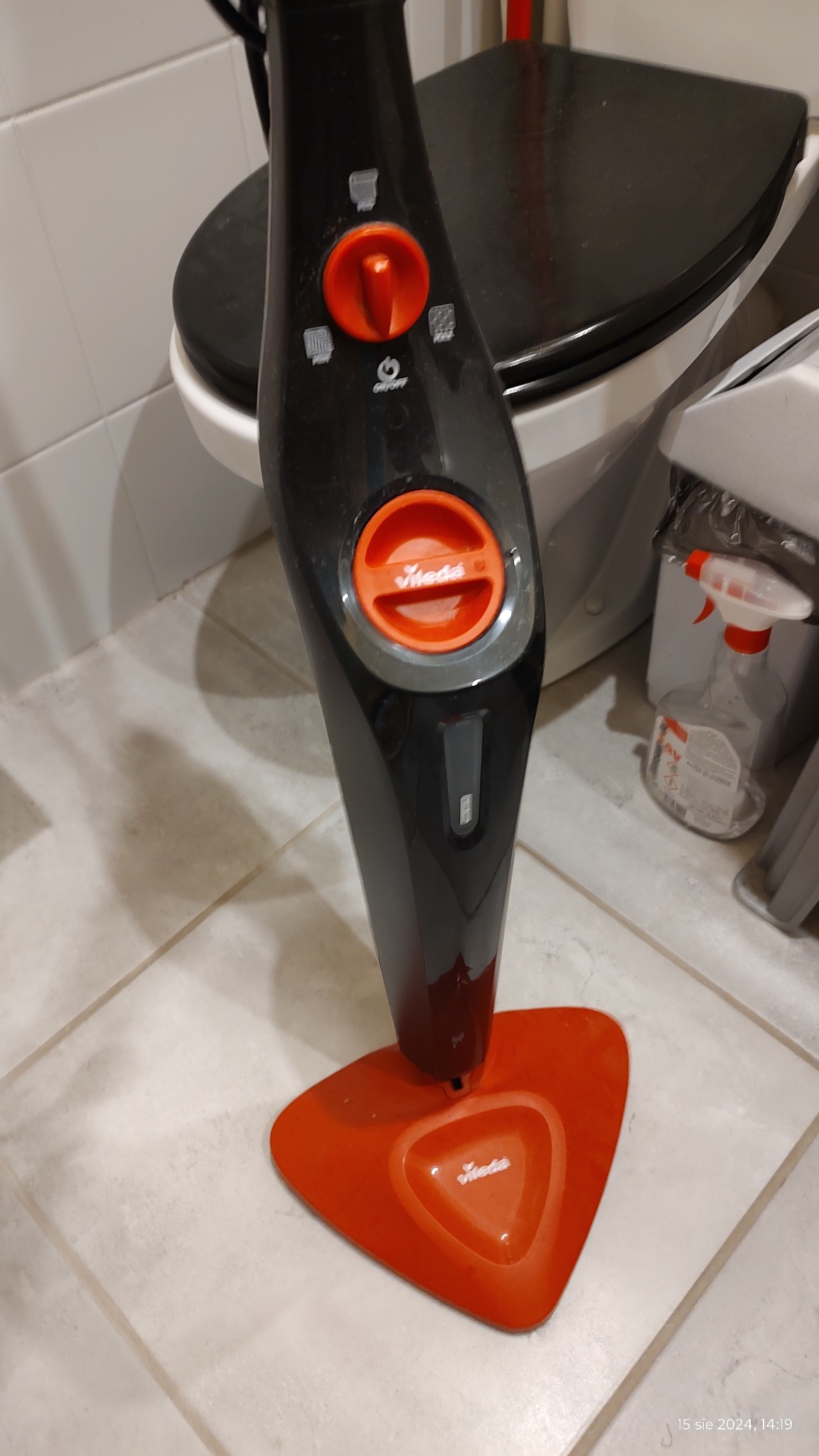 Czarny mop parowy Vileda z pomarańczowymi akcentami stoi na jasnych płytkach podłogowych w łazience, obok toalety i butelki ze spryskiwaczem.