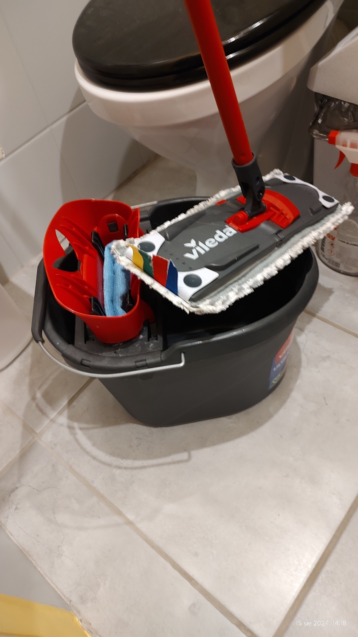 Mop Vileda z czerwoną rączką i szarą nakładką w szarym wiadrze z wyciskaczem, obok toaleta i butelka ze spryskiwaczem.