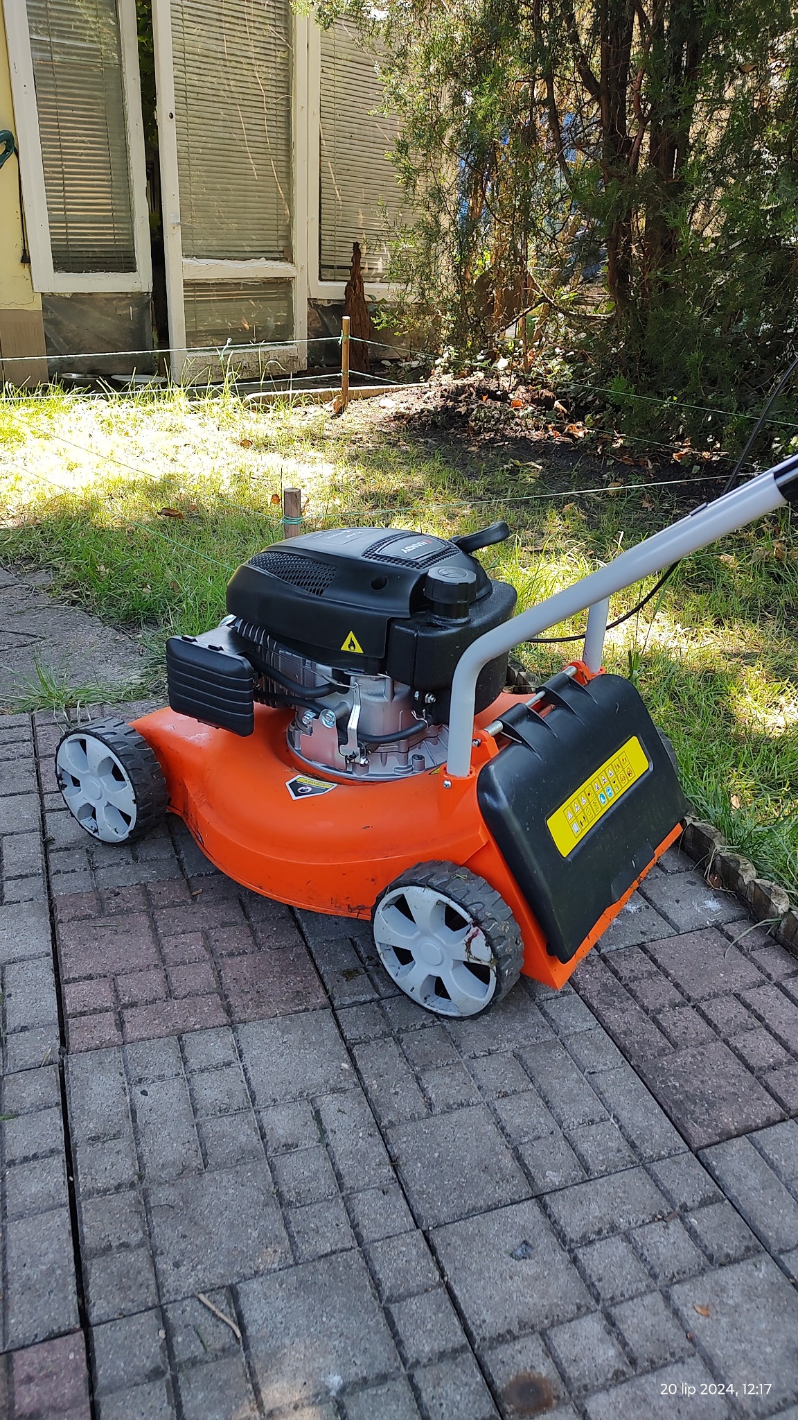 Pomarańczowa kosiarka spalinowa, częściowo widoczna na kostce brukowej, trawa i budynek w tle.