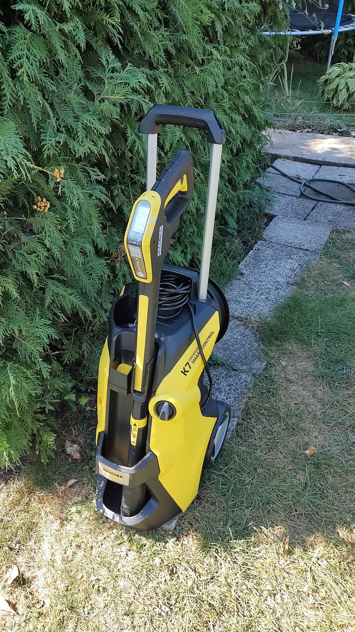 Żółto-czarna myjka ciśnieniowa Karcher K7 Smart Control, stojąca na trawie obok zielonego żywopłotu i kamiennej ścieżki.