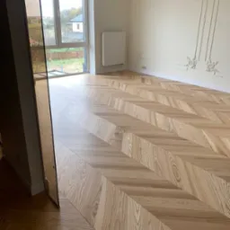 Nowo ułożony parkiet w jodełkę klasyczną, jasne drewno, widok na okno i dekoracyjne sztukaterie na ścianie.