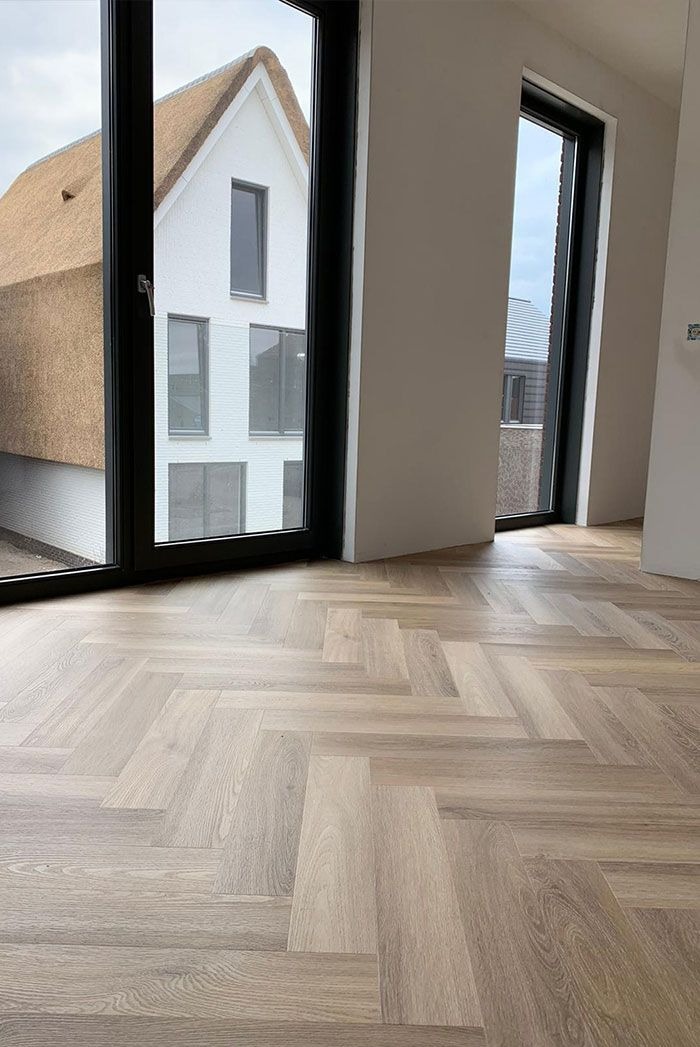 Jasny parkiet ułożony we wzór jodełki w nowym wnętrzu, widok przez okno na dom z charakterystycznym strzechą.