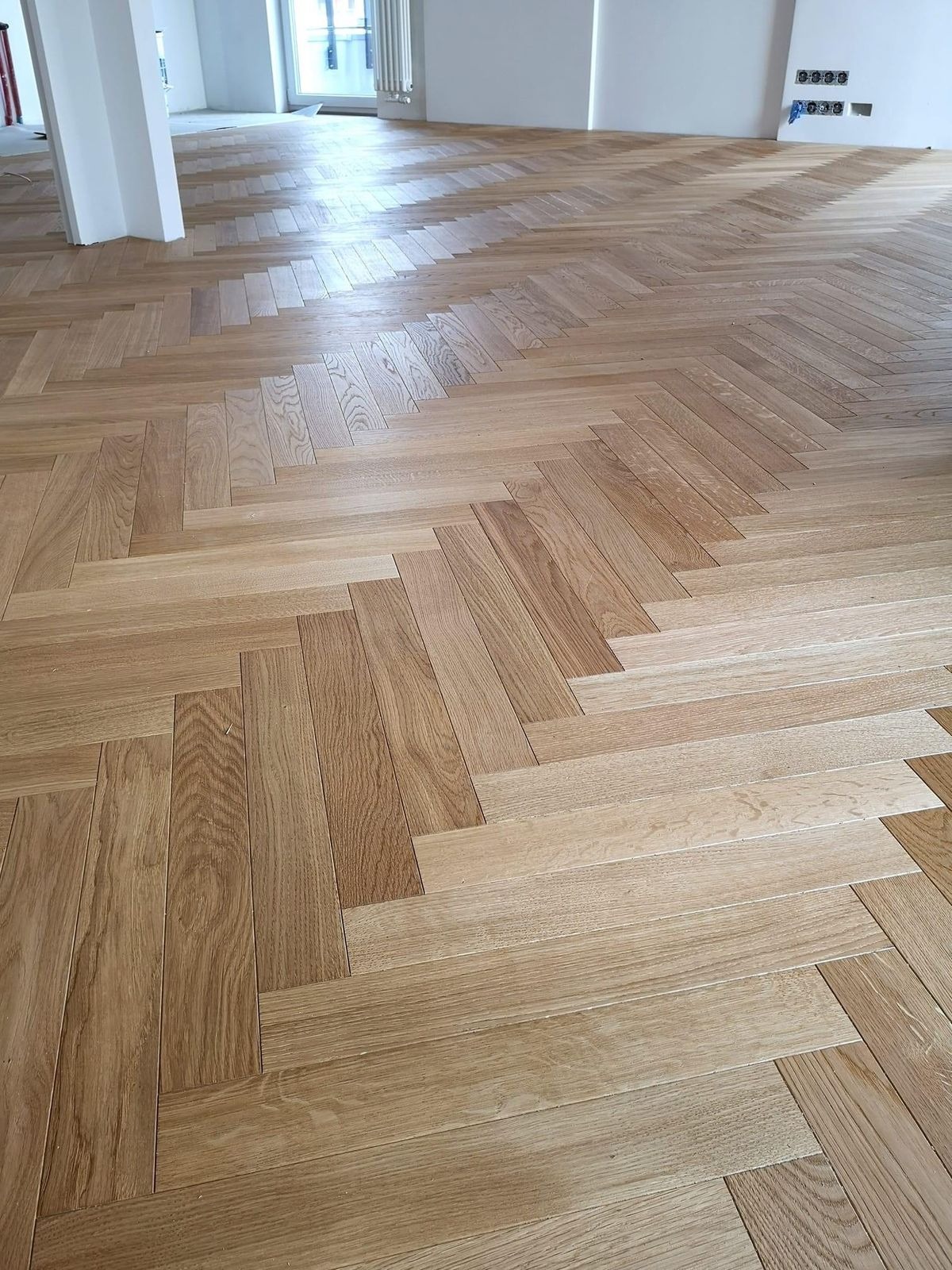 Jasnobeżowa podłoga z parkietu ułożonego we wzór jodełki francuskiej w przestronnym, jasnym pomieszczeniu w trakcie remontu, widoczne niedokończone fragmenty i instalacja elektryczna na ścianie.