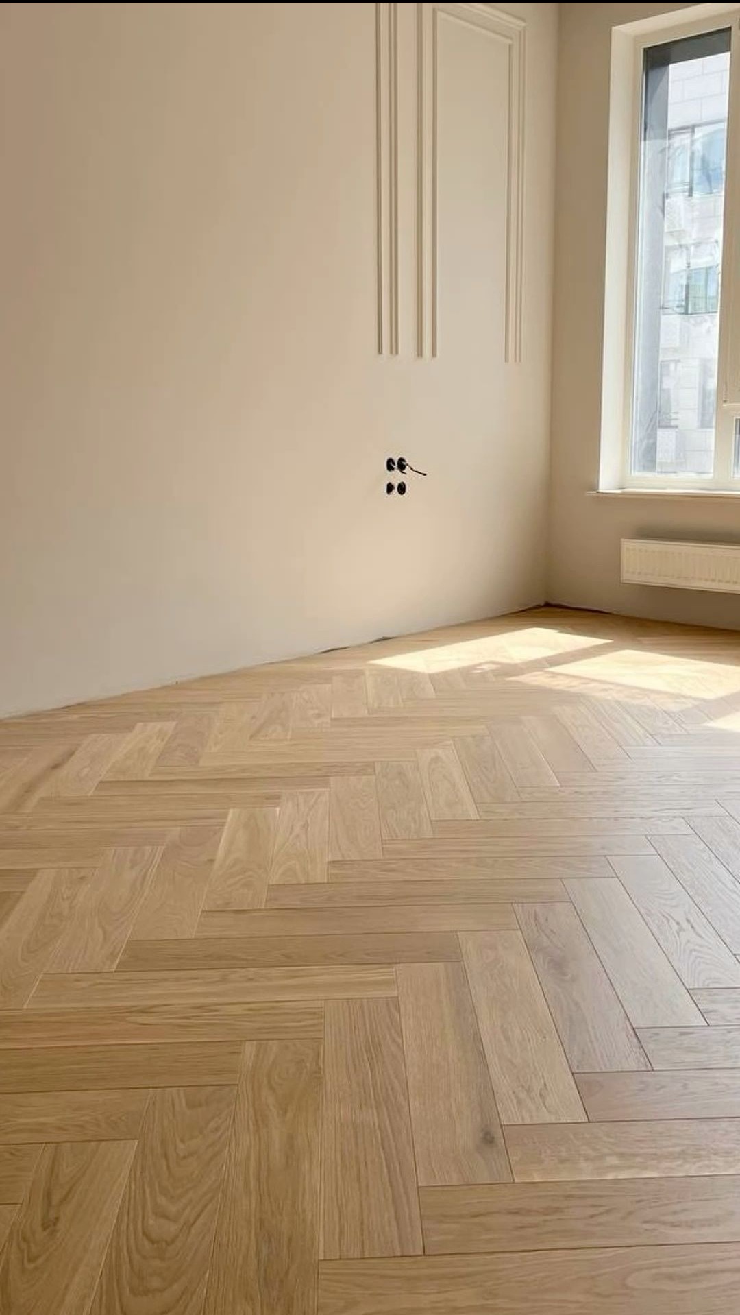 Jasny parkiet ułożony we wzór jodełki francuskiej w nowym pomieszczeniu z dekoracyjnymi listwami na ścianie i dużym oknem.