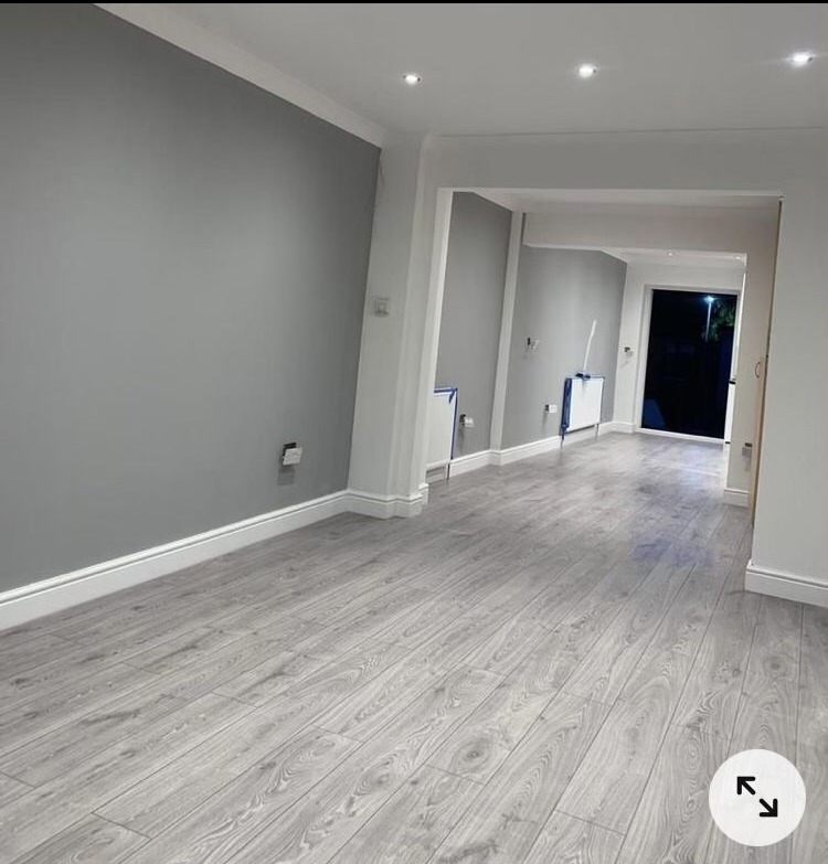 Pokój po remoncie z szarymi ścianami i jasną, drewnopodobną podłogą, widoczne listwy przypodłogowe i przejście do kolejnego pomieszczenia.