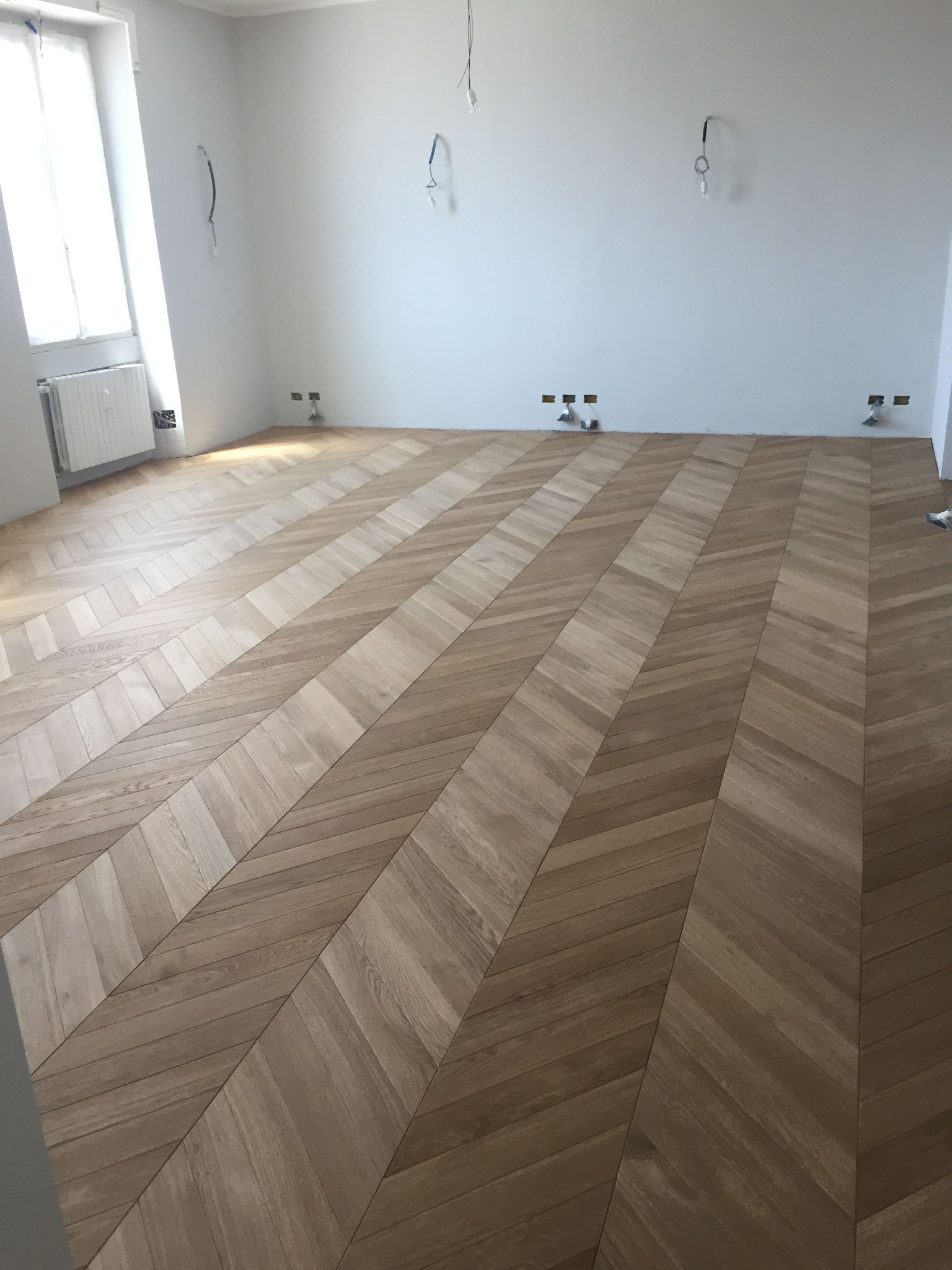 Nowo ułożony parkiet w jodełkę w jasnym, pustym pokoju z białymi ścianami i przygotowanymi punktami oświetleniowymi.