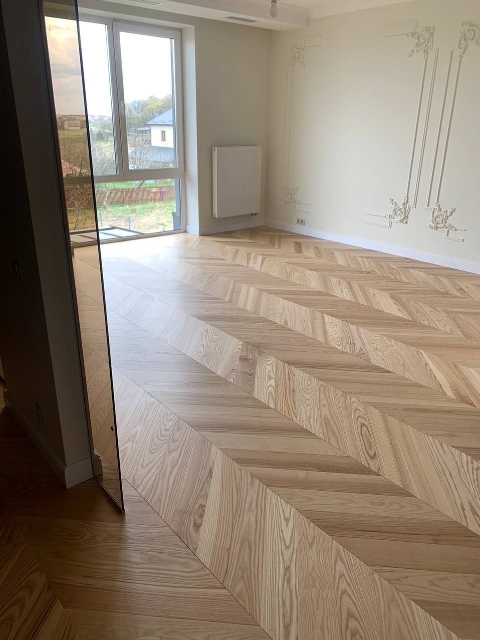 Nowo ułożony parkiet w jodełkę klasyczną, jasne drewno, widok na okno i dekoracyjne sztukaterie na ścianie.