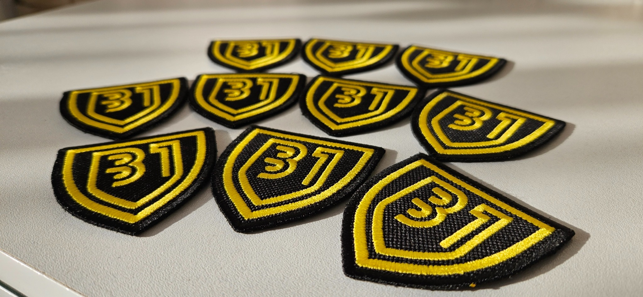 Grupowe ujęcie dziesięciu czarnych naszywek w kształcie tarczy z wyhaftowanym żółtym obrysem i numerem '31', leżących na gładkiej, jasnej powierzchni.