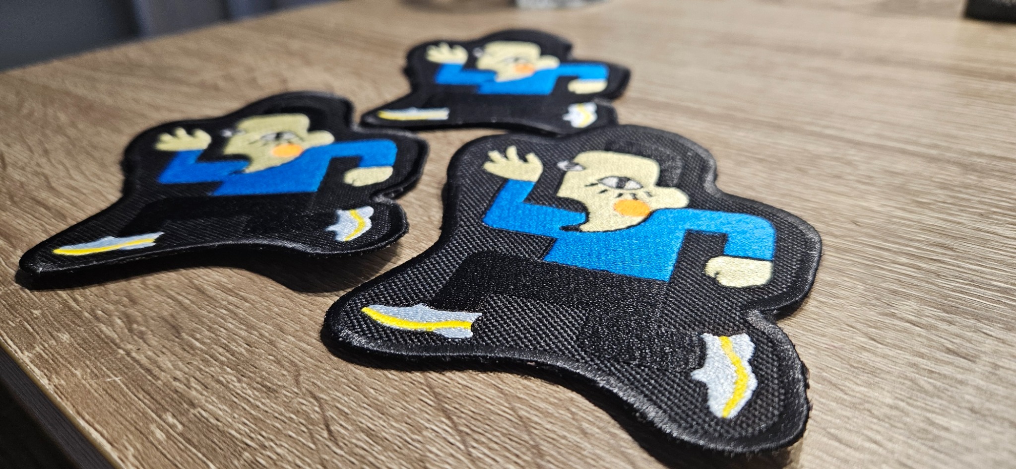 Trzy czarne naszywki z haftowanym, stylizowanym wizerunkiem postaci w niebieskiej koszuli, leżące na drewnianej powierzchni.