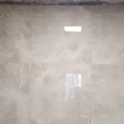 Ściany łazienki wyłożone szarymi, matowymi płytkami imitującymi beton, z widocznym otworem na oświetlenie i fragmentem drewnianej futryny drzwi po lewej stronie, na górze ściany widoczne ślady po...