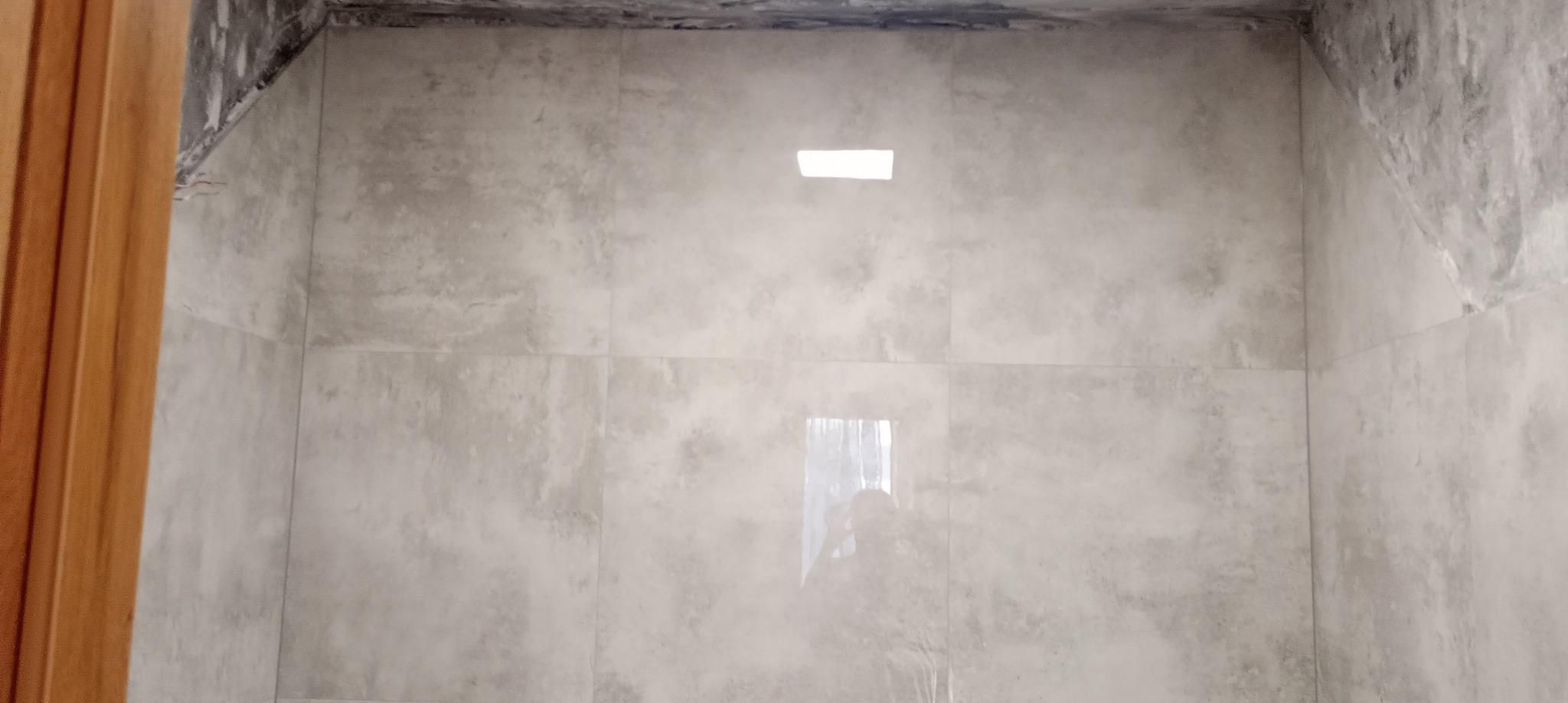 Ściany łazienki wyłożone szarymi, matowymi płytkami imitującymi beton, z widocznym otworem na oświetlenie i fragmentem drewnianej futryny drzwi po lewej stronie, na górze ściany widoczne ślady po...