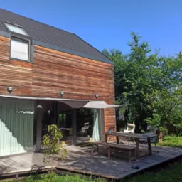 Nowoczesny dom z drewnianą elewacją i tarasem, na którym stoi drewniany stół z ławkami i kwiatami w doniczce, zacieniony rozpiętą tkaniną.