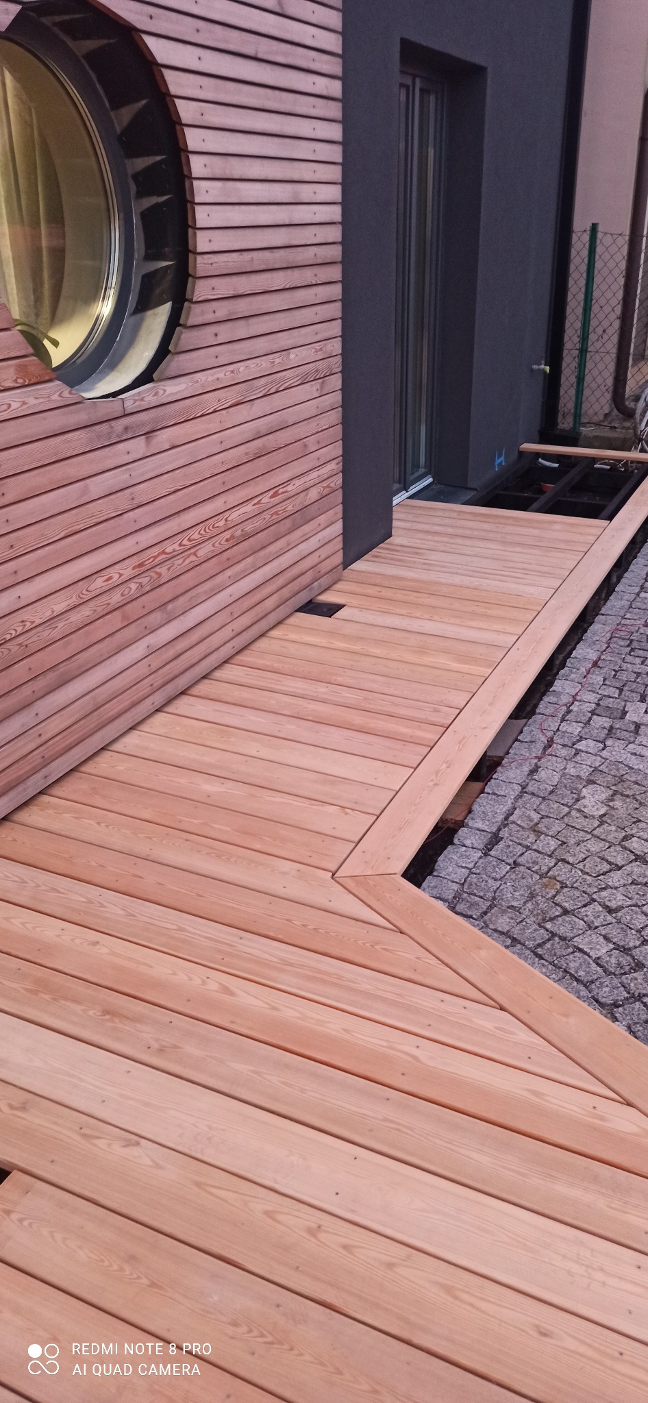 Montaż jasnego tarasu z desek drewnianych przy nowoczesnym domu z okrągłym oknem, widoczne elementy konstrukcyjne pod tarasem.