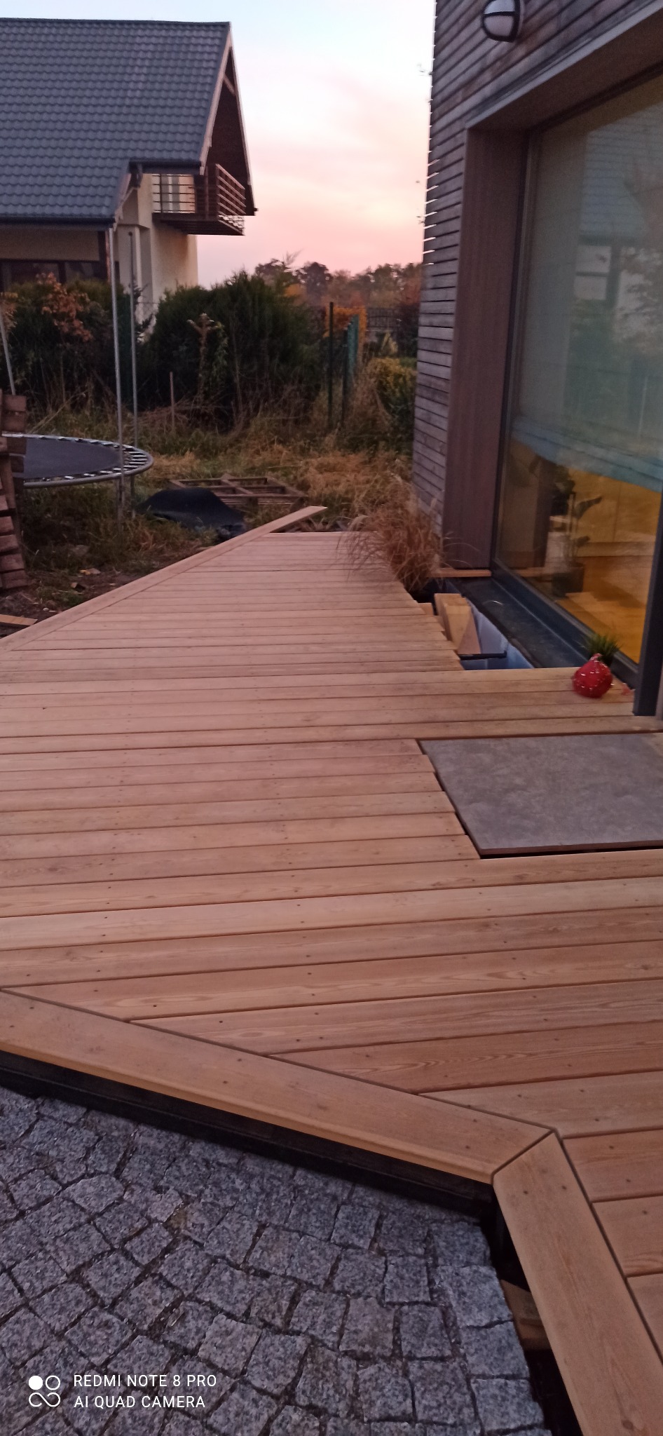 Nowoczesny taras z jasnego drewna, częściowo ukończony, z widoczną kostką brukową i elementami konstrukcyjnymi domu w tle.