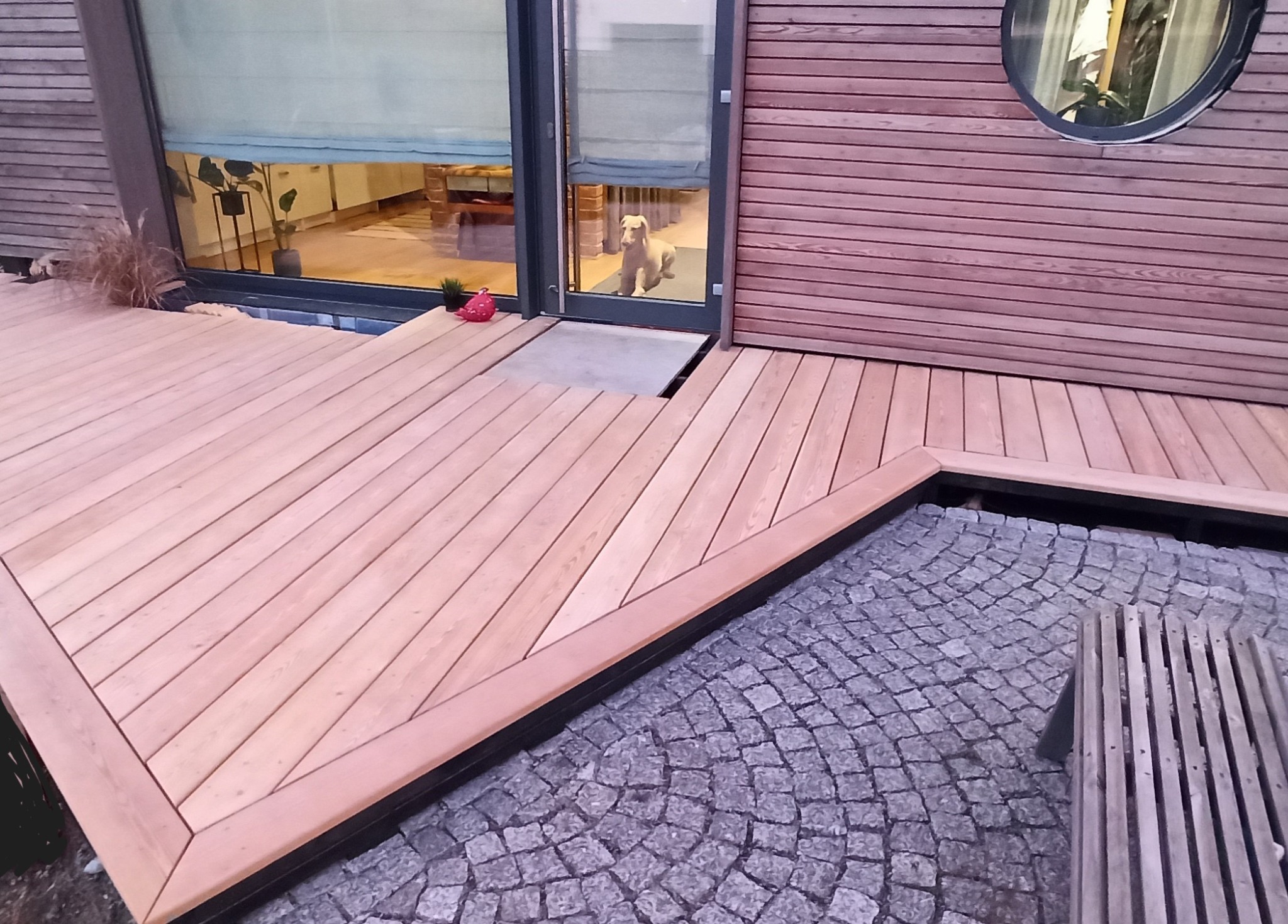 Jasne deski tarasu ułożone pod kątem prostym, zintegrowane z elewacją domu wykończoną drewnem, widoczna kostka brukowa i pies w oknie.