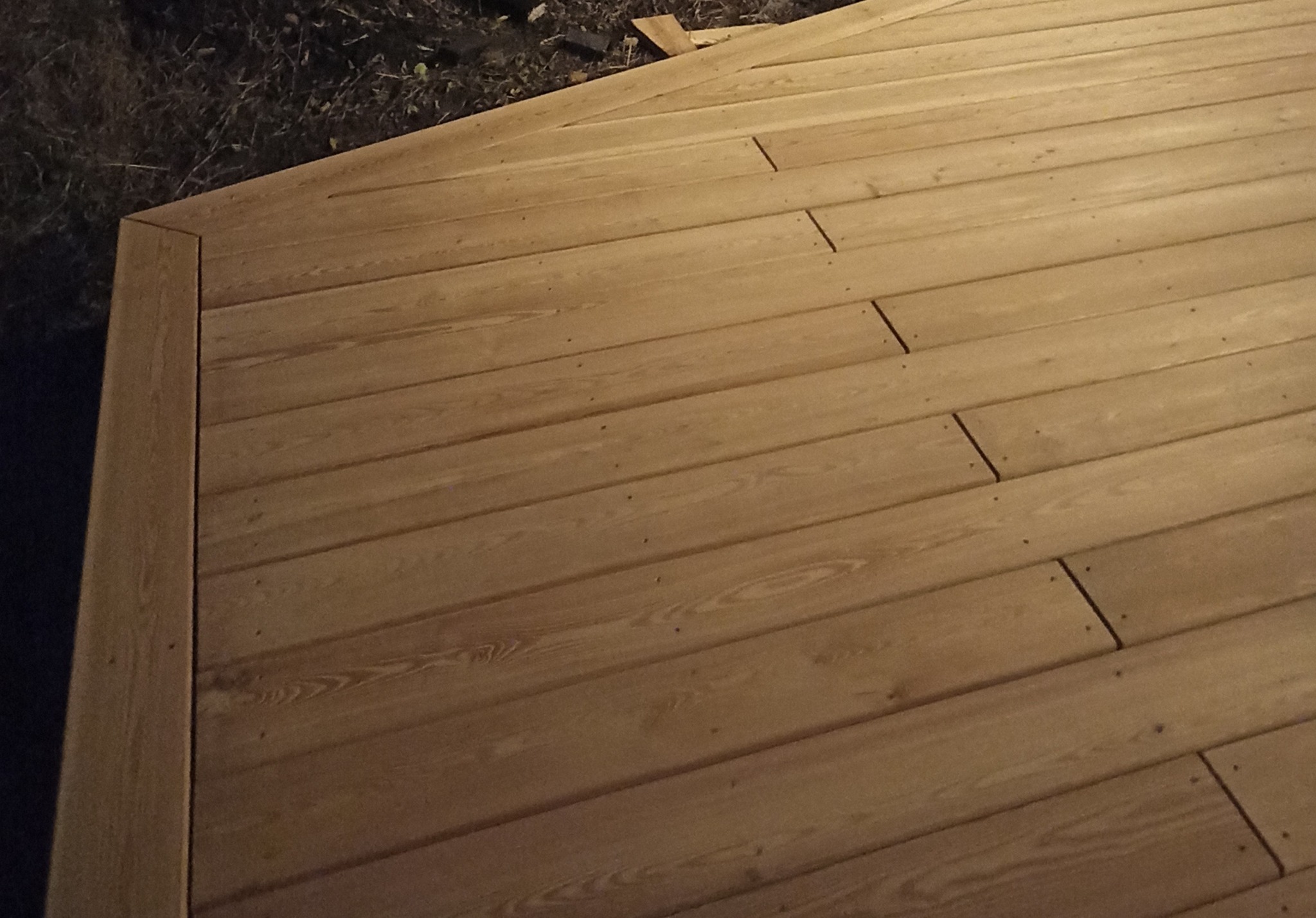 Narożnik nowej, jasnej, drewnianej podłogi tarasowej, widoczne wkręty mocujące deski, tło z ciemną roślinnością.