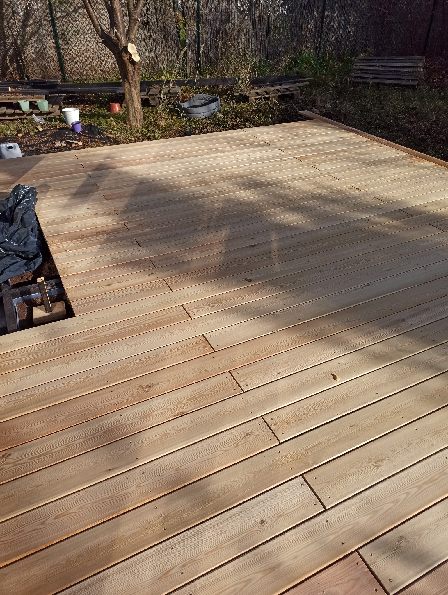 Montaż desek na tarasie drewnianym; widoczny fragment niedokończonej podkonstrukcji z czarną folią, deski w jasnym odcieniu, cień drzewa na powierzchni.