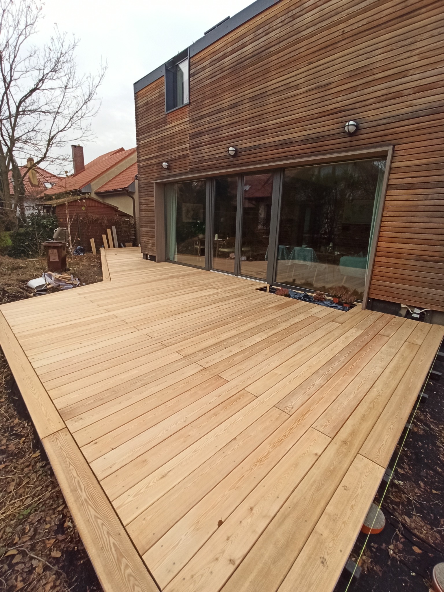 Jasna, drewniana deska tarasowa przylegająca do budynku z drewnianą elewacją, widok na ogród i sąsiednie domy w tle.