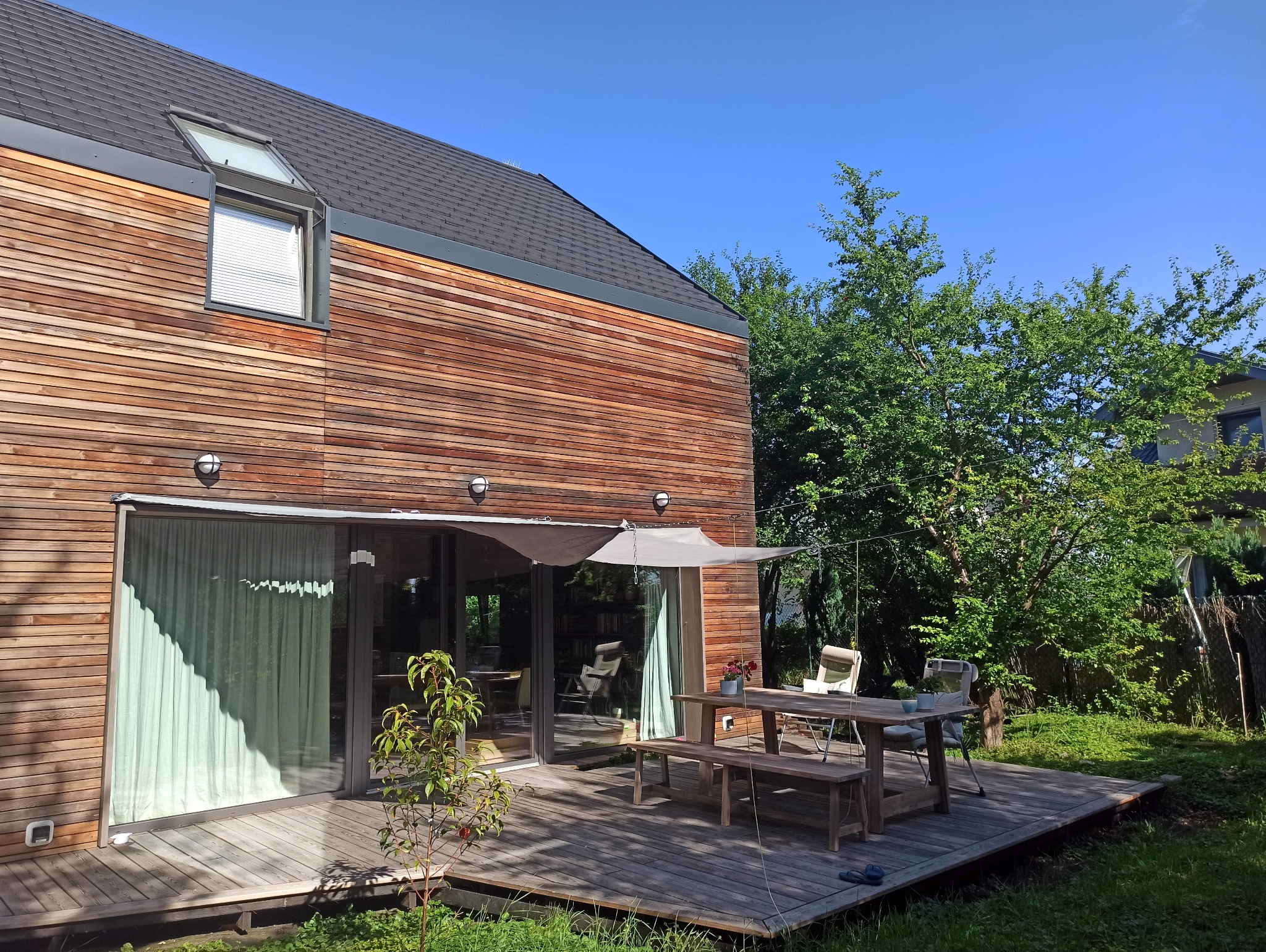 Nowoczesny dom z drewnianą elewacją i tarasem, na którym stoi drewniany stół z ławkami i kwiatami w doniczce, zacieniony rozpiętą tkaniną.