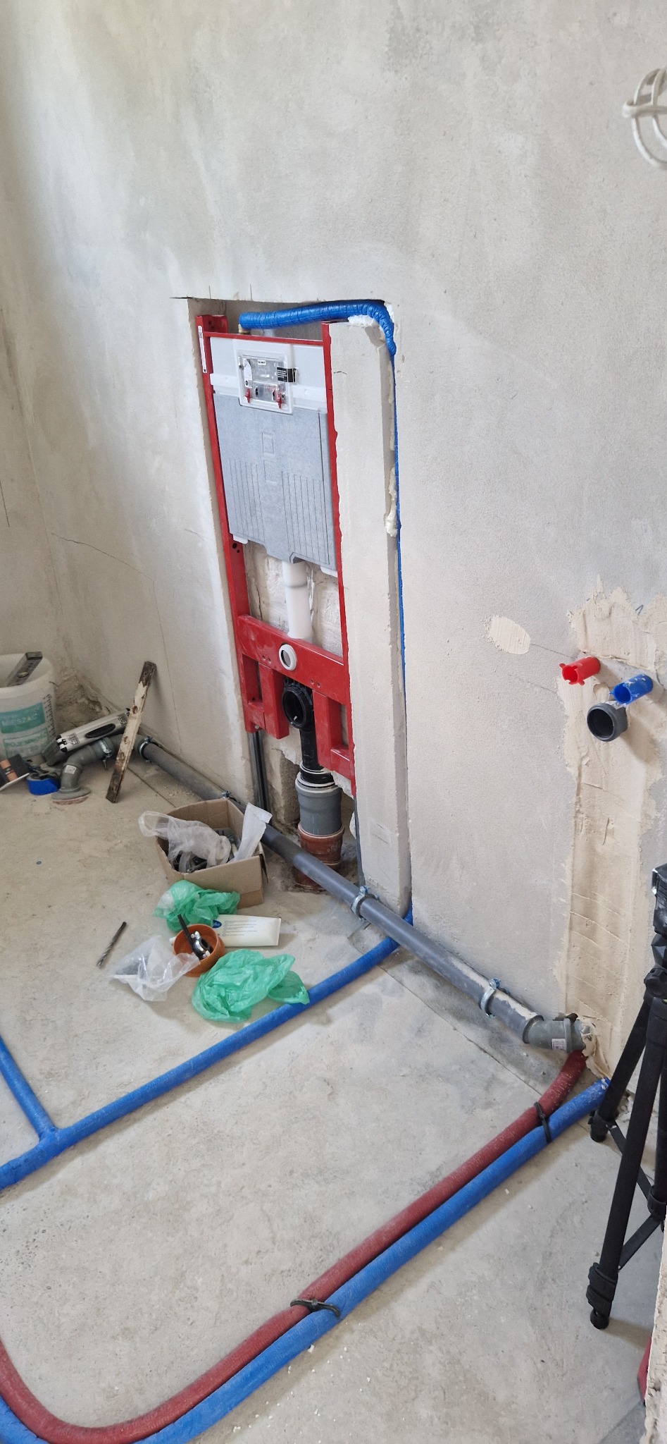 Wnęka w surowej ścianie z zamontowanym czerwonym stelażem podtynkowym TECE do WC, widoczne podłączenia wodno-kanalizacyjne, rury i narzędzia w trakcie montażu.