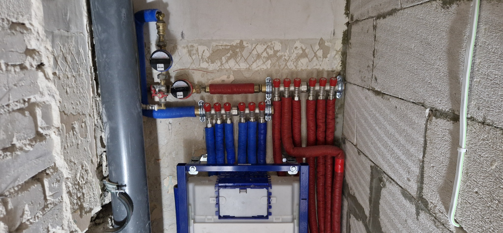 Rozdzielacze ciepłej i zimnej wody zamontowane w bloku, z widocznymi zaworami w kolorze czerwonym do regulacji dopływu wody, instalacja wykonana na surowych ścianach z betonu komórkowego.