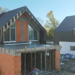 Budowa nowoczesnego domu murowanego z płaskim dachem i oknami trójkątnymi w szczycie, widoczne szalunki i podpory stropu, w tle drugi budynek z blachy na rąbek.