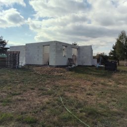 M KRUK POLSKA - Wznoszenie ścian domu z betonu komórkowego. Widoczne palety z bloczkami i drabina. Budowa w toku, na zewnątrz, w otoczeniu drzew i trawy.