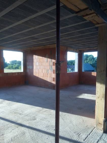 Wnętrze surowego domu murowanego w trakcie budowy, widoczne ściany z czerwonej cegły, betonowa podłoga i okna z widokiem na zieleń, podpora stropu na pierwszym planie.