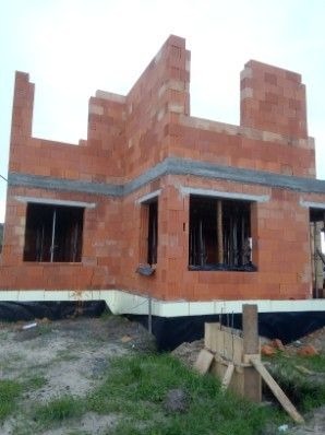 Surowy stan budynku z czerwonej cegły, widoczne otwory okienne i fragment ściany szczytowej, fundament zabezpieczony izolacją, drewniane szalunki w tle.