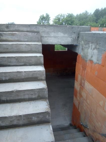 Surowe betonowe schody prowadzące do piwnicy w trakcie budowy, z widocznymi fragmentami murowanych ścian z czerwonej cegły i betonową nadprożem.